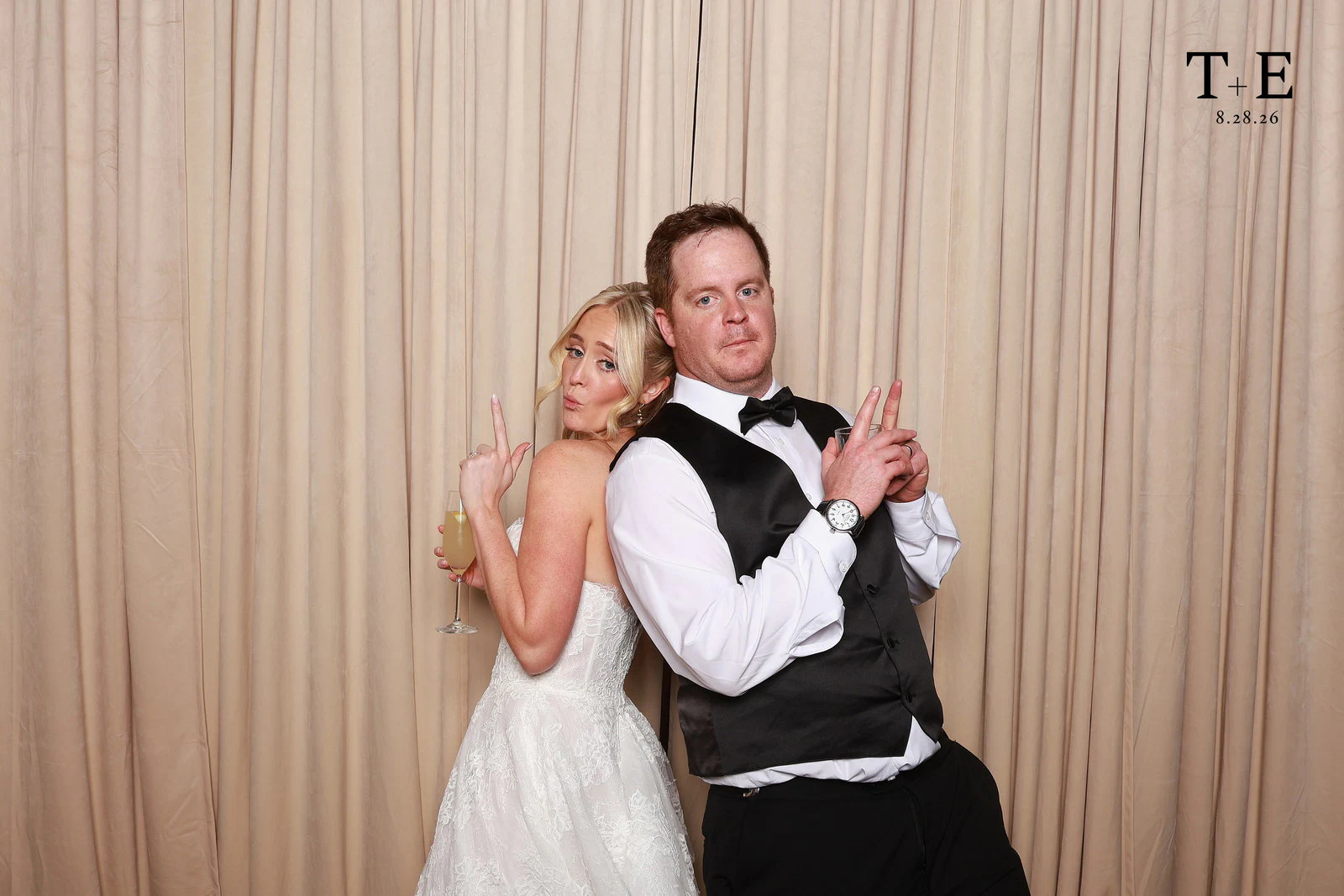 Tara in her white gown and Ethan in a black waistcoat and bow tie, back to back with finger guns, in front of the beige velvet at Tara + Ethan’s wedding