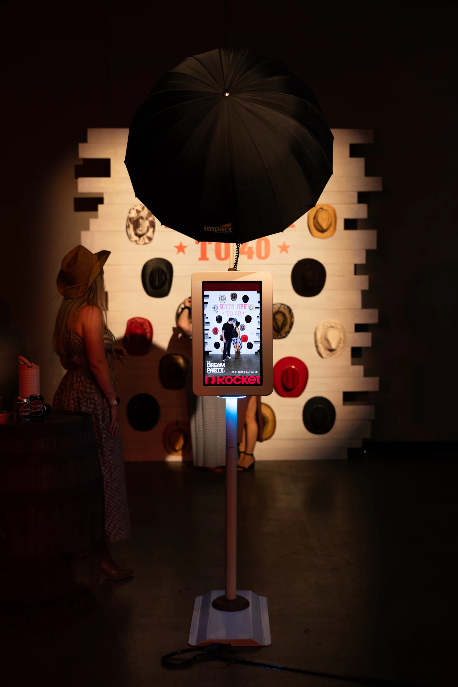 The booth screen and umbrella light in front of the Hats Off to 40 wall, a guest in a cowboy hat nearby at The Dream Party: Rocket at 40