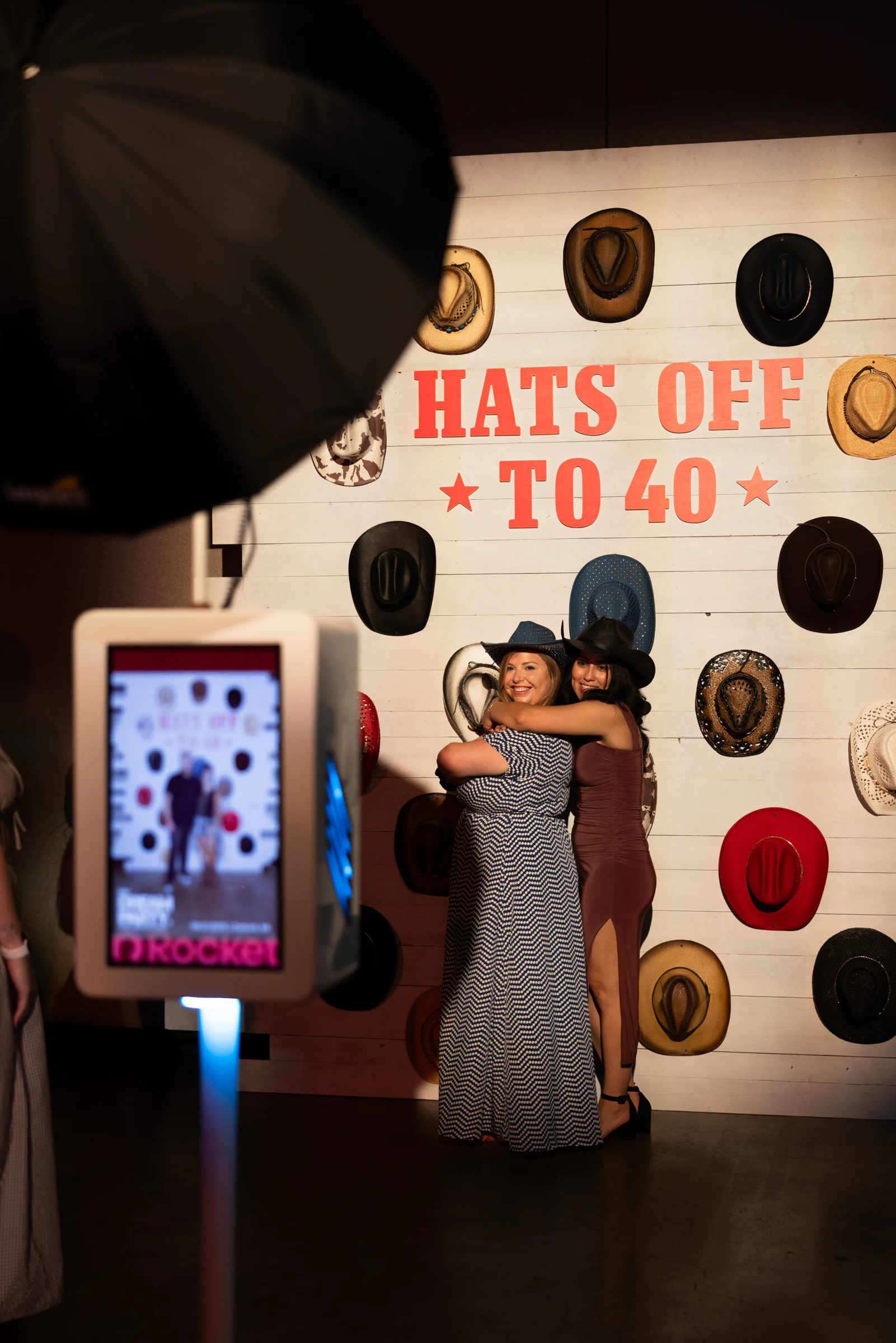 Two guests in cowboy hats hugging at the Hats Off to 40 wall, the booth screen in the foreground at The Dream Party: Rocket at 40