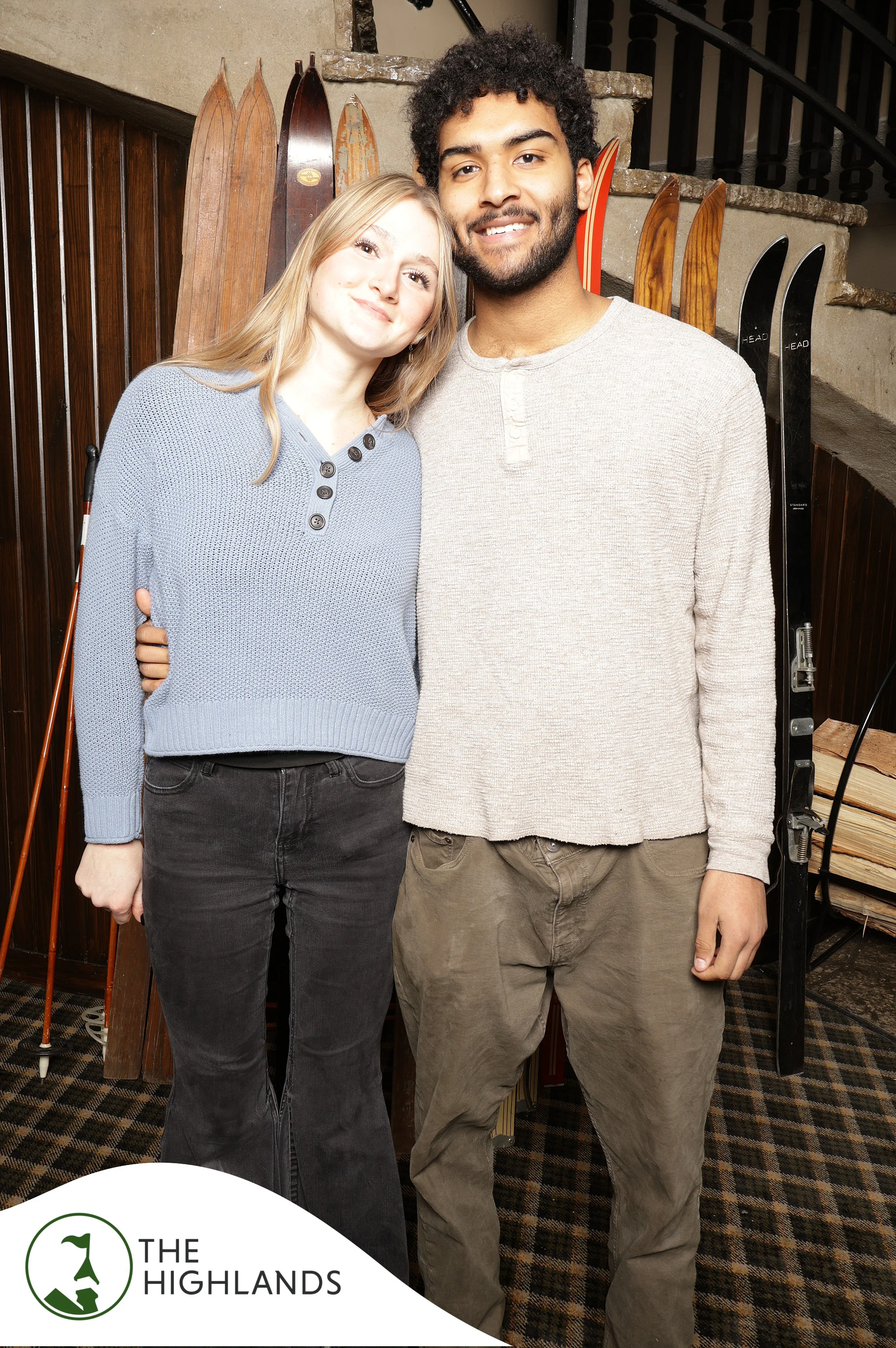 Couple in soft sweaters standing close in front of the vintage skis at The Highlands 61st Anniversary