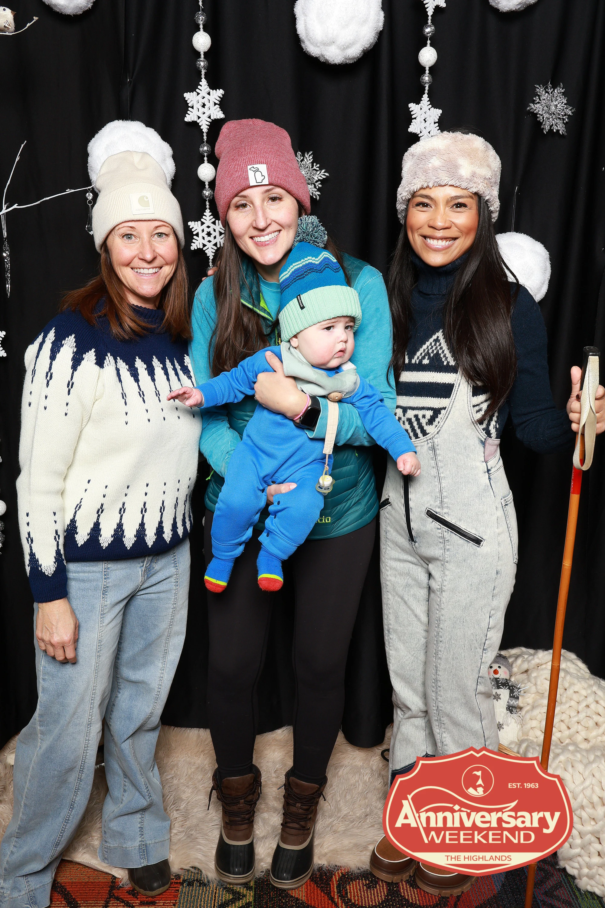 Three women in ski sweaters and beanies, one holding a baby in a blue snowsuit, in front of the snowflake backdrop at The Highlands Anniversary Weekend