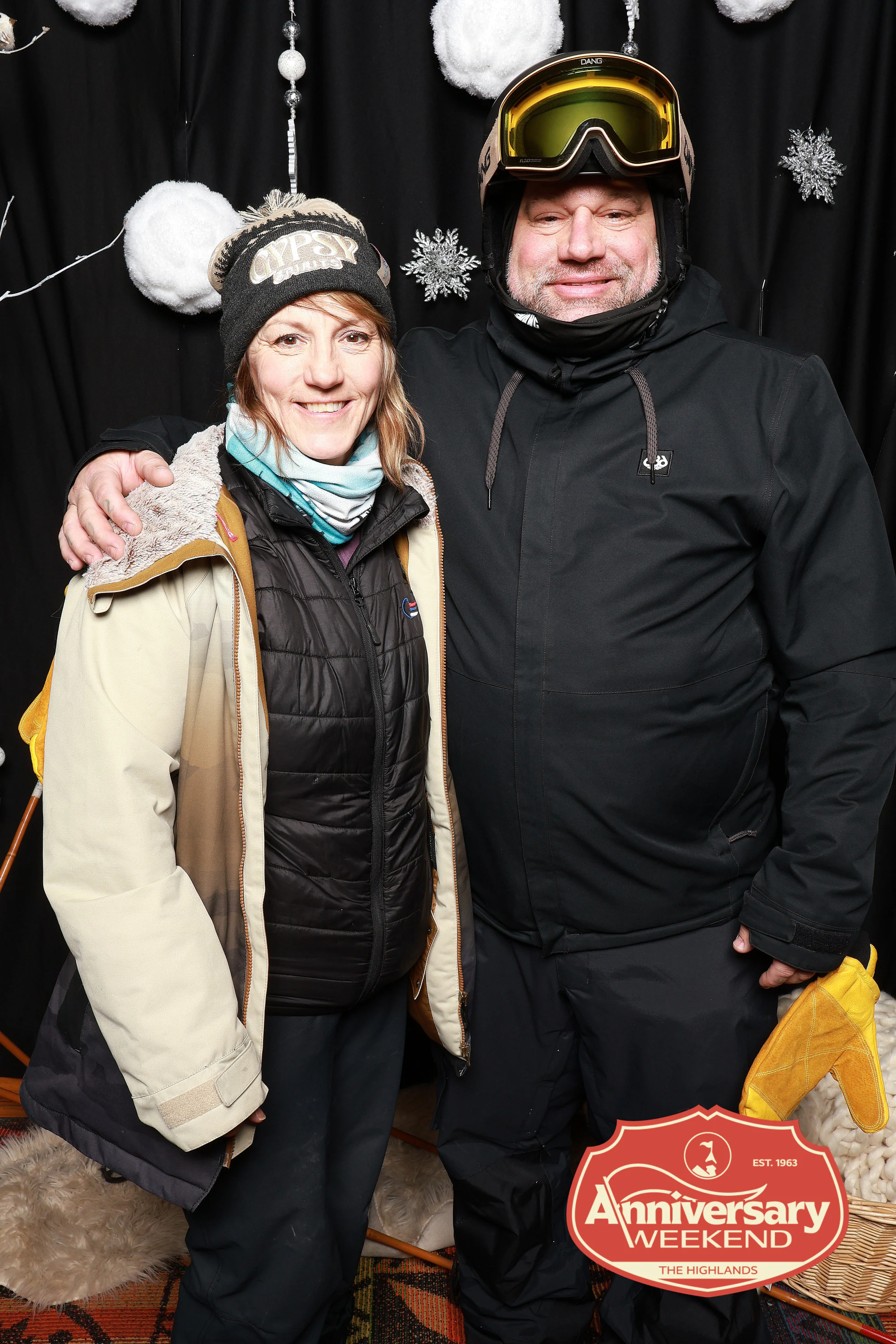 The couple in ski kit smiling in front of the snowflake backdrop at The Highlands Anniversary Weekend