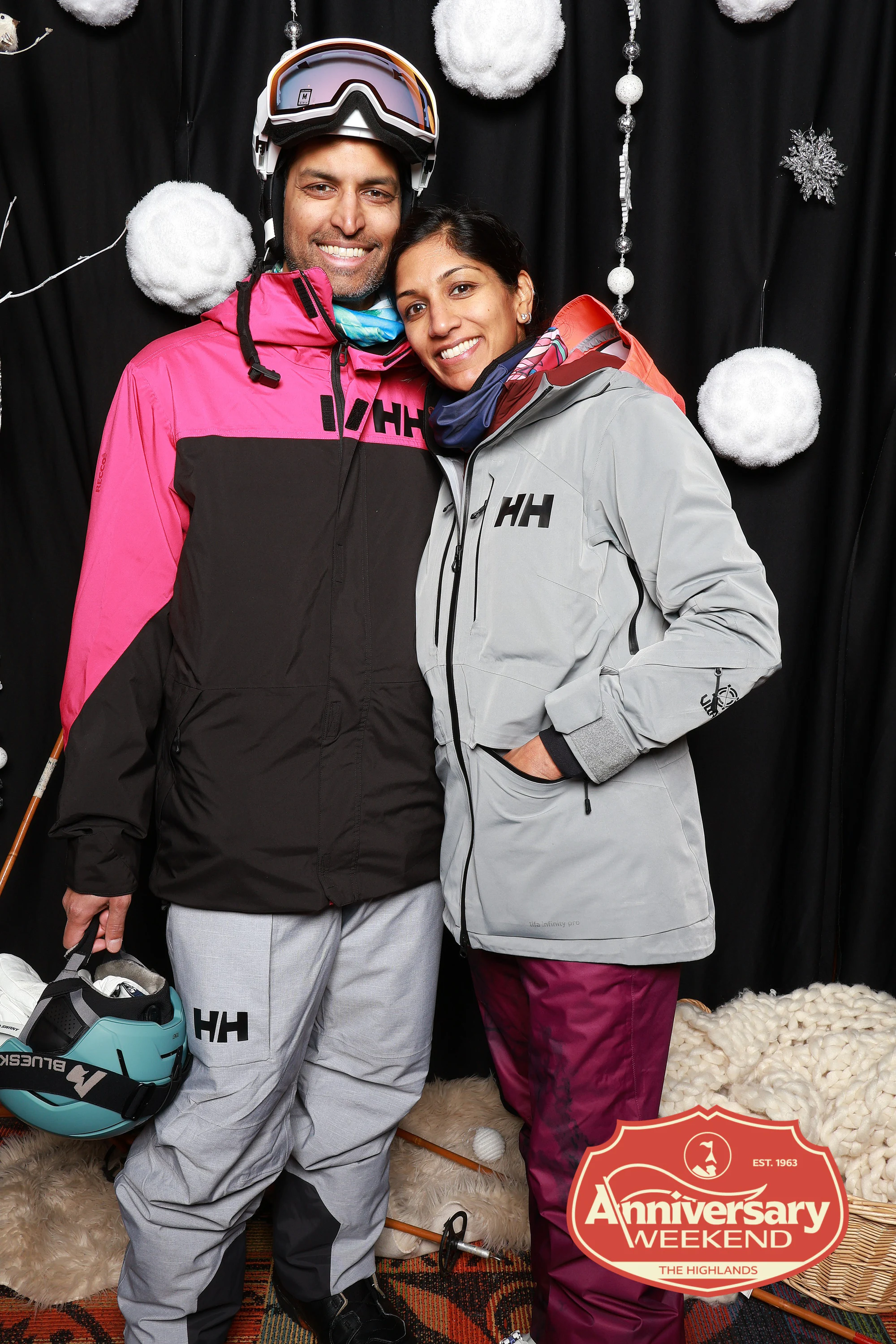 Couple in ski jackets in front of the snowflake backdrop, his goggles up on his helmet at The Highlands Anniversary Weekend