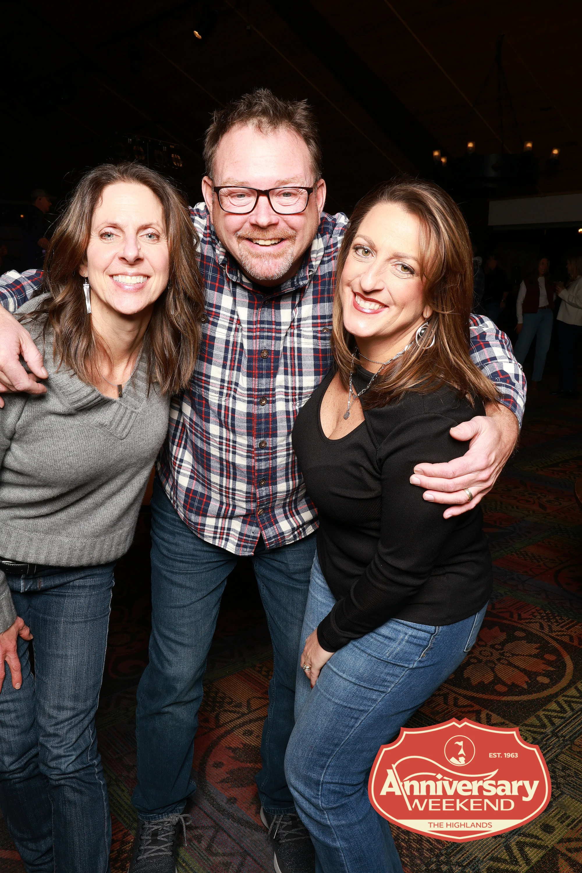 Man in a plaid shirt with an arm around two friends at the evening party at The Highlands Anniversary Weekend