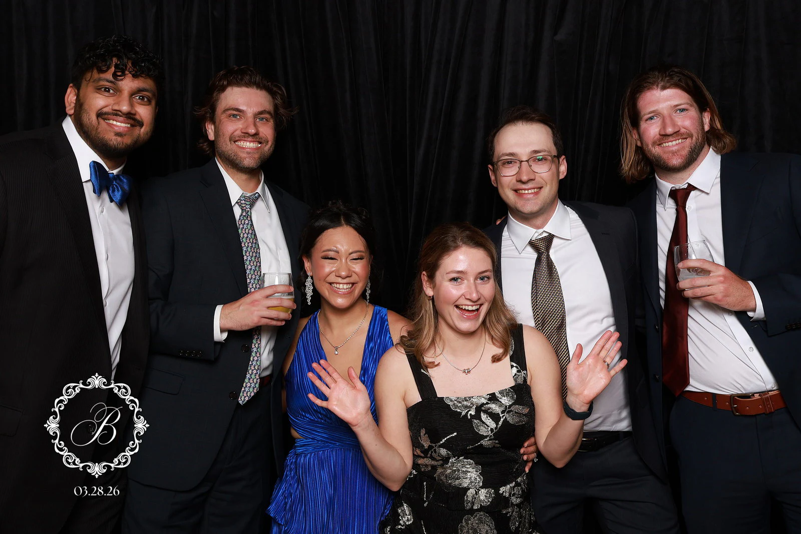 Four men in suits standing behind two women waving at the camera at Tianna + Krishna
