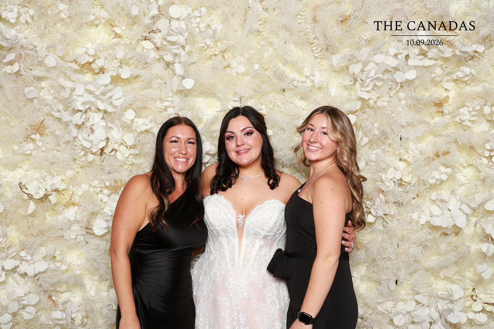 The bride between two friends in black dresses, all three smiling in front of the Champagne Dream flower wall at Tristin + Emma