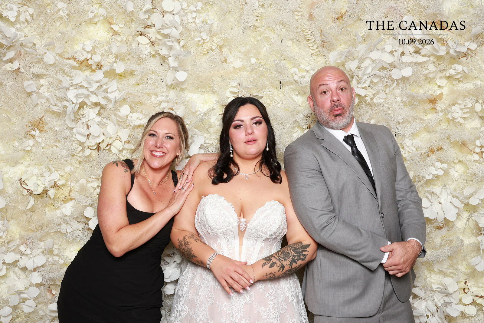 The bride with a woman in black leaning on her shoulder and a man in a gray suit puckering up for the camera at Tristin + Emma