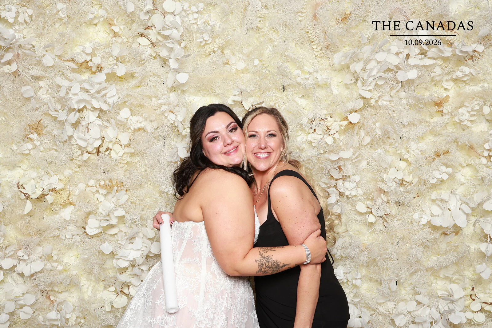 The bride and a friend in black hugging cheek to cheek in front of the flower wall at Tristin + Emma