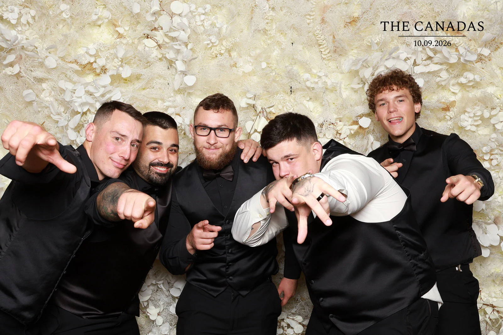 The groom and four groomsmen in black vests leaning in and pointing at the camera at Tristin + Emma