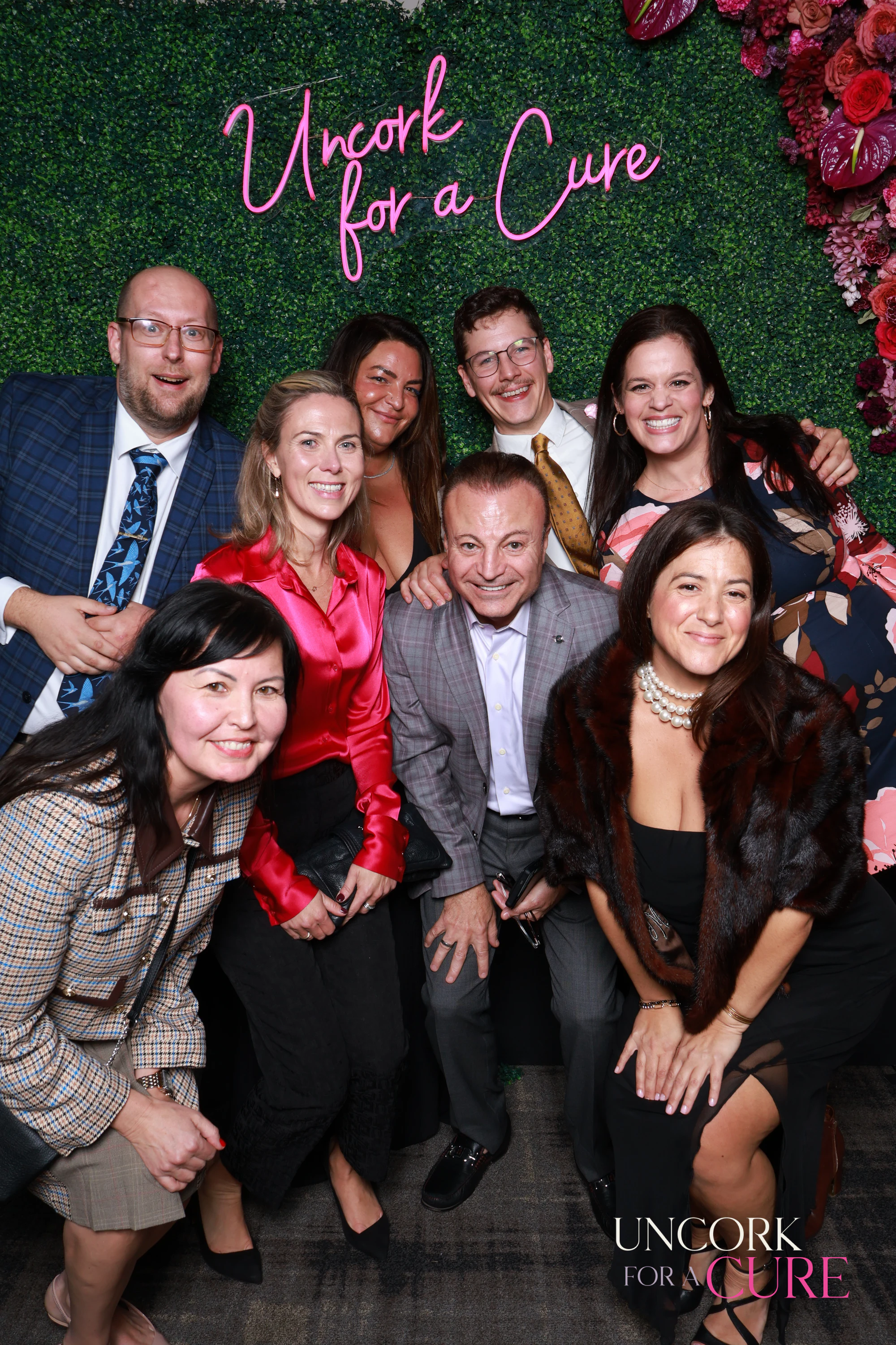 Ten guests under the Uncork for a Cure sign, some crouched in front at Uncork for a Cure 2026