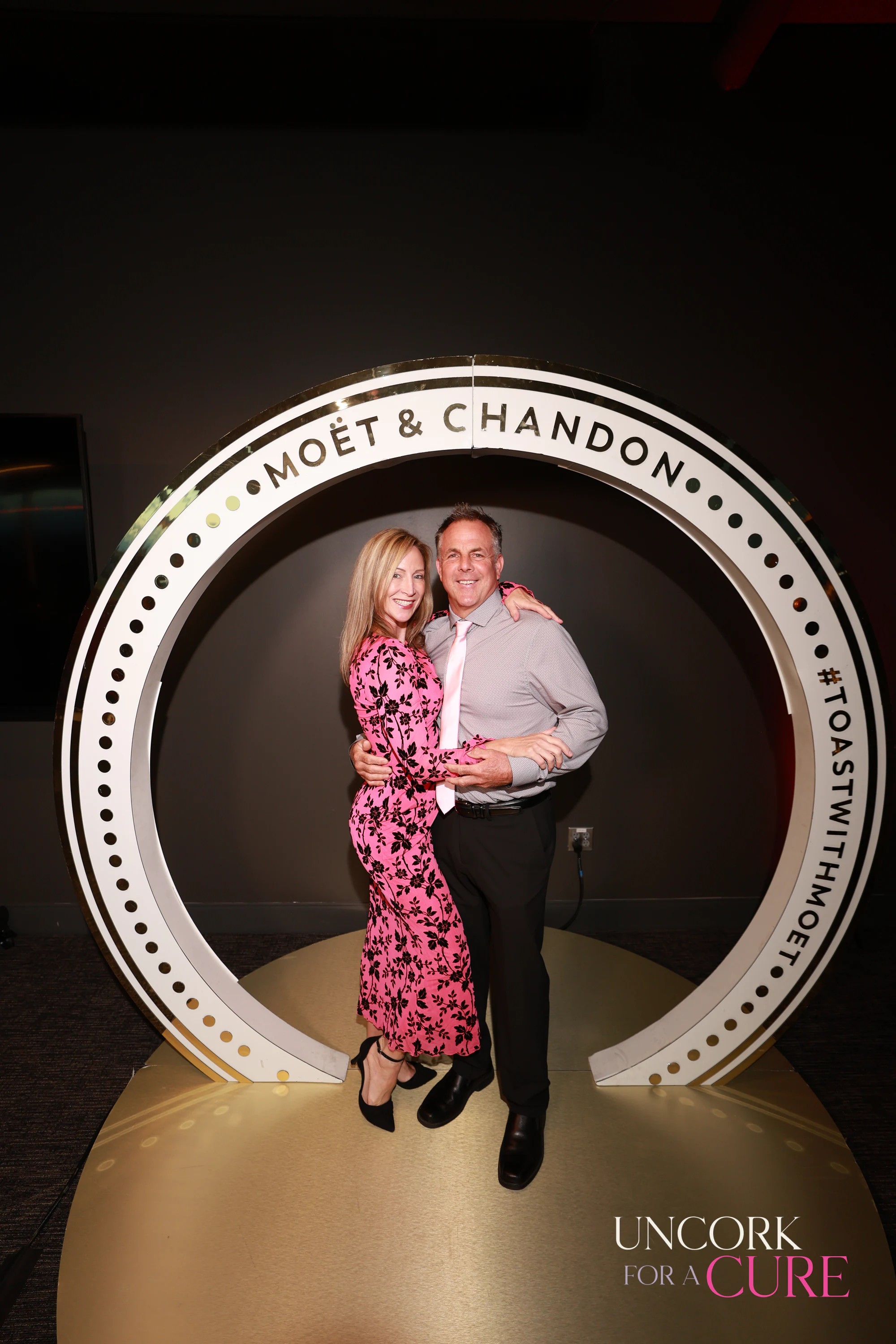 A couple standing in the Moët & Chandon ring, she in a pink floral dress at Uncork for a Cure 2026