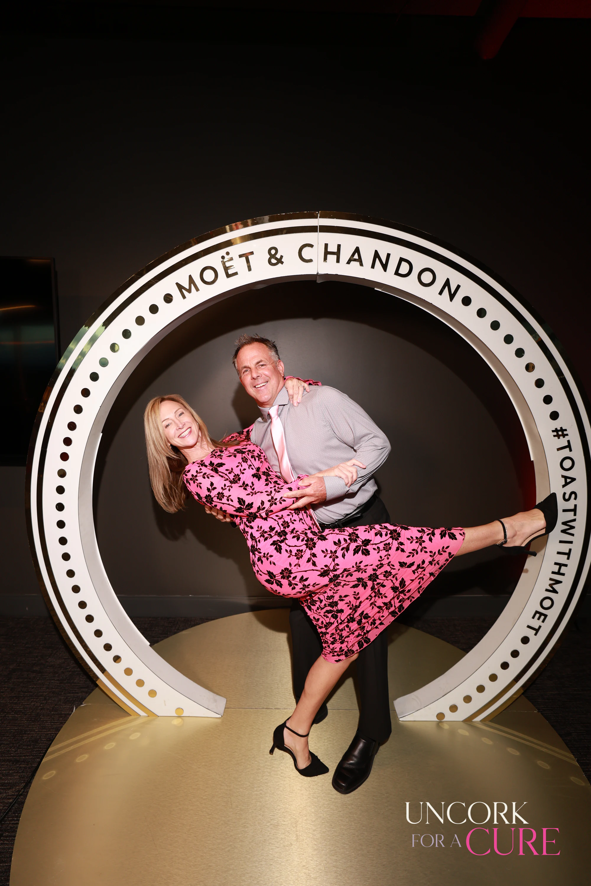 The woman in pink floral kicking up a leg as the man holds her in the Moët & Chandon ring at Uncork for a Cure 2026