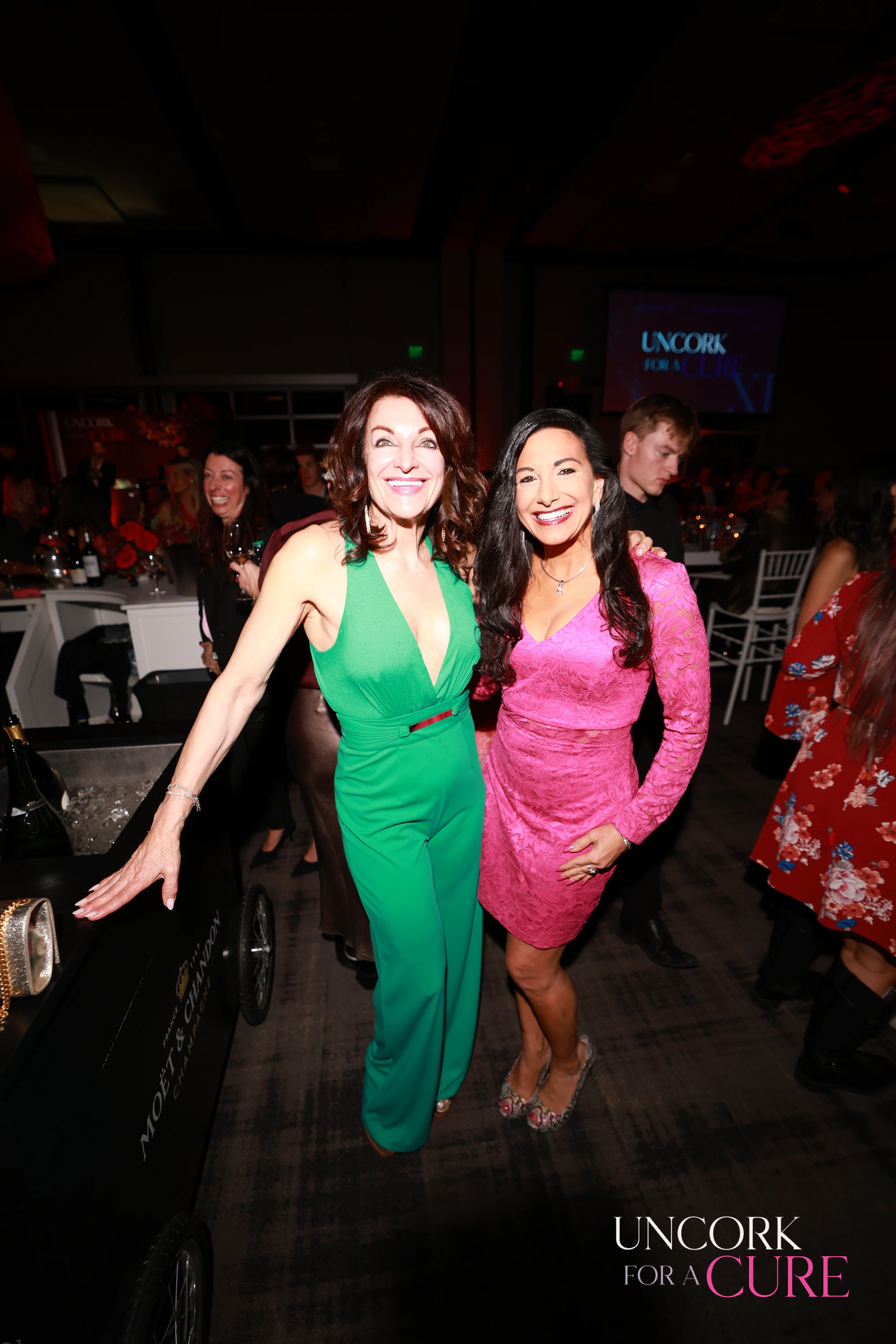 The two women in green and pink posing, one with an arm out at Uncork for a Cure 2026