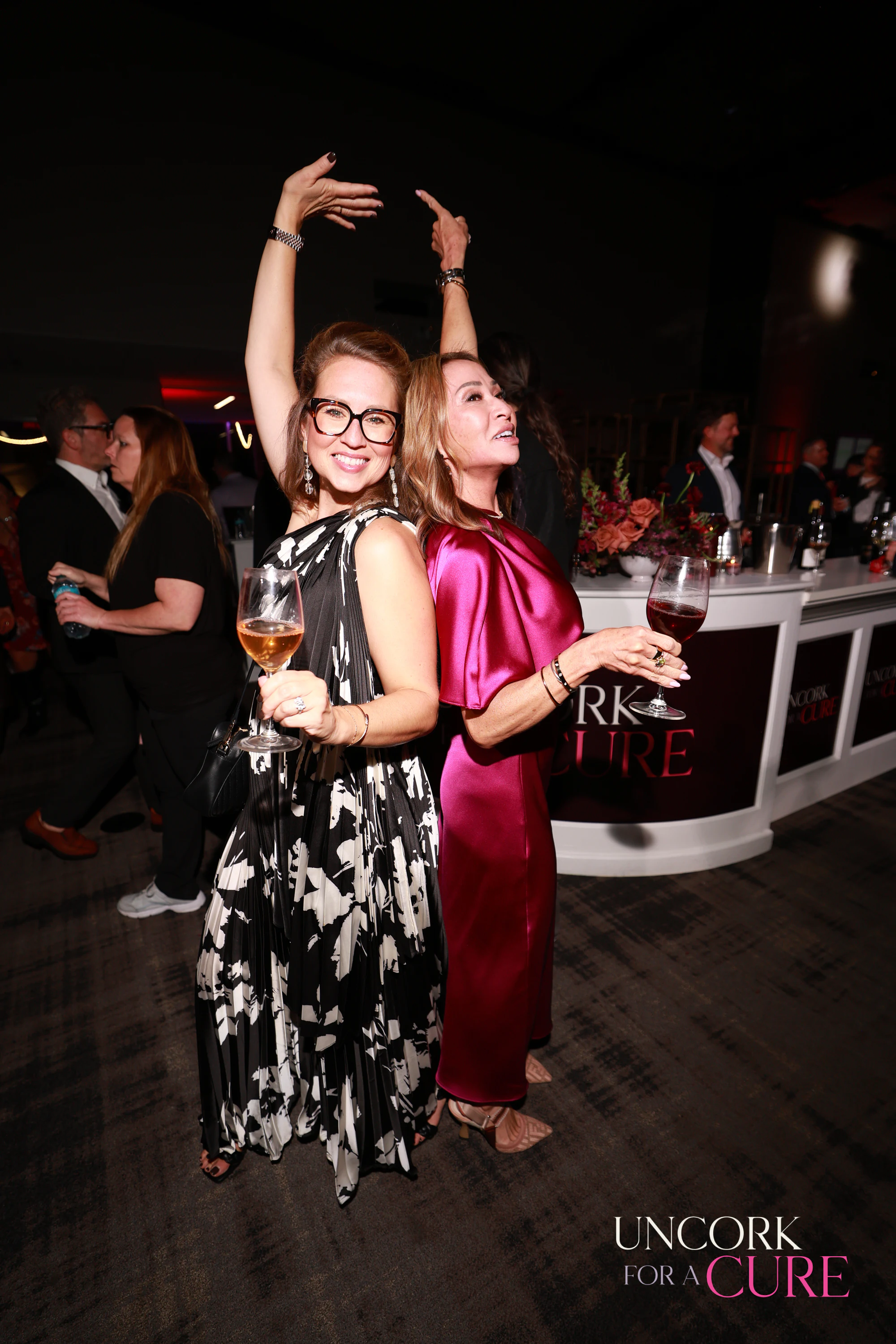 Two women dancing, one with her arms curved over her head at Uncork for a Cure 2026